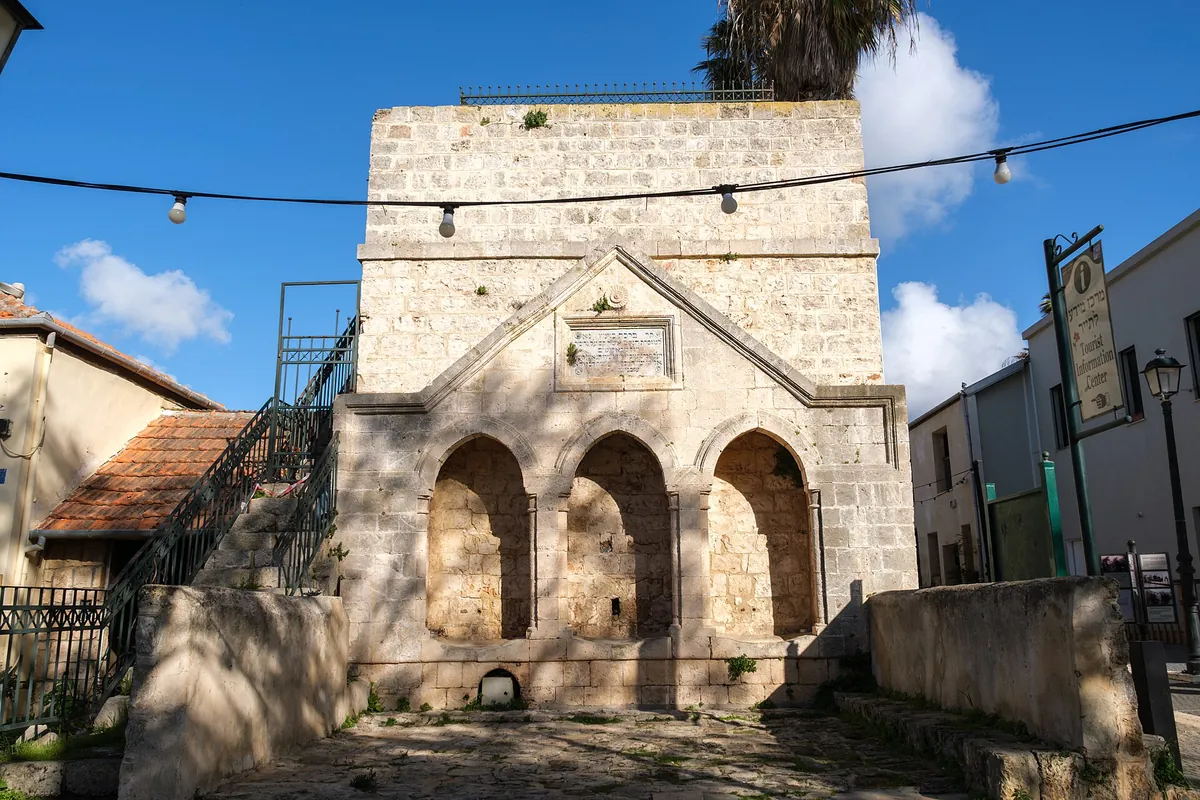 Stone water-reservoir building with three arched niches and an inscription plaque, beside a tourist information sign on HaMeyasdim Street