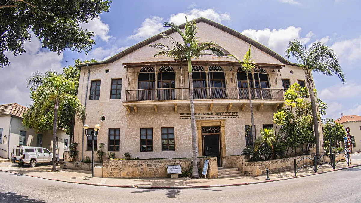The First Aliyah Museum: a two-storey stone building with a wooden balcony, framed by palm trees
