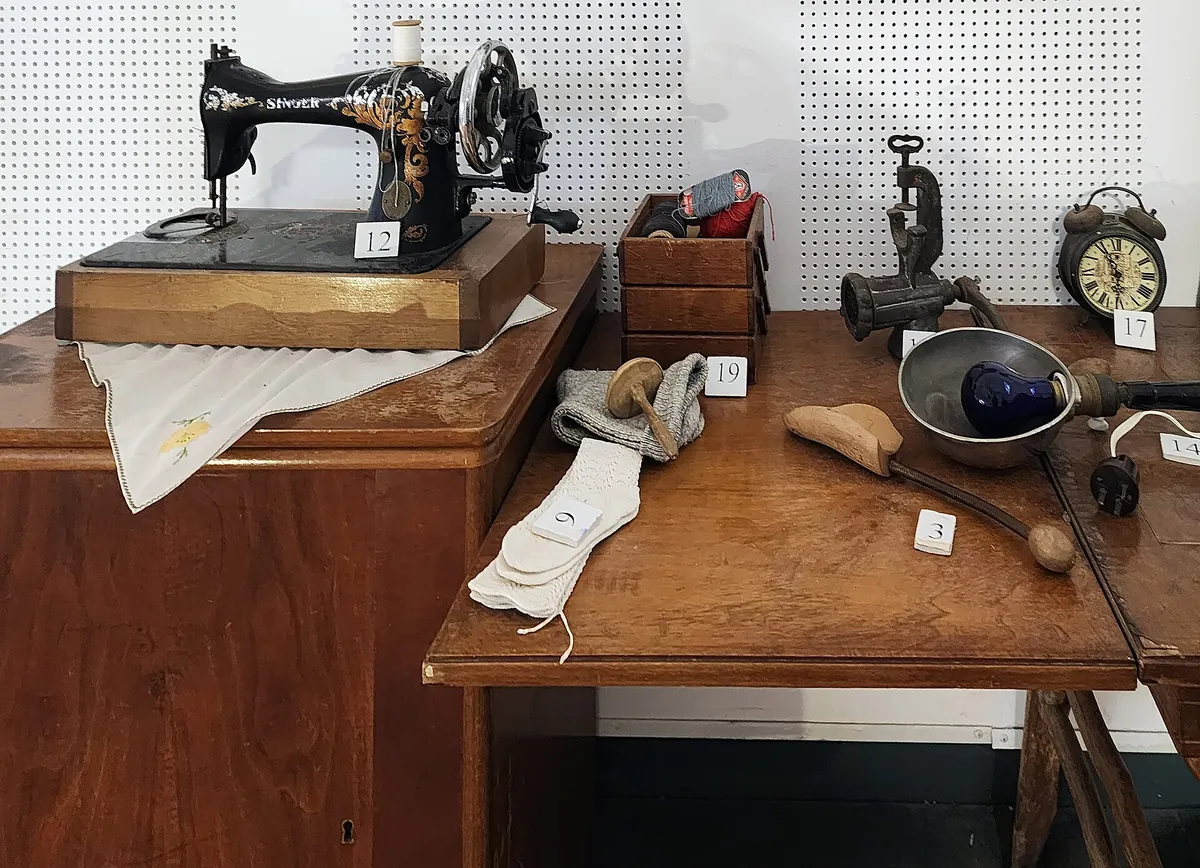 Exhibit inside the First Aliyah Museum: an old sewing machine, a clock and household tools on wooden furniture